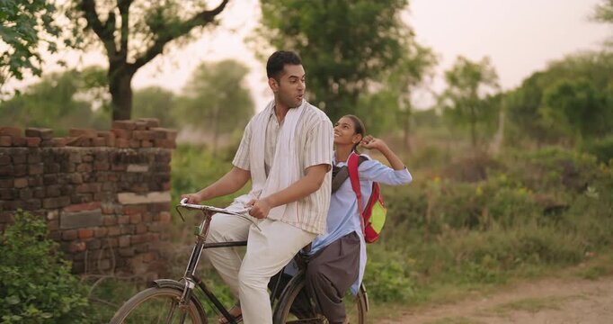 Indian happy young adult dad talk speak drop school to teen girl child rural area place road path way. Smiling female wear uniform with bag sitting on cycle back seat enjoy ride have fun joy desi life
