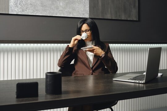 A real estate agent in a brown suit sits at a desk, sipping coffee from a small cup in a contemporary office. Her laptop is open, but she decided to make a coffe-break