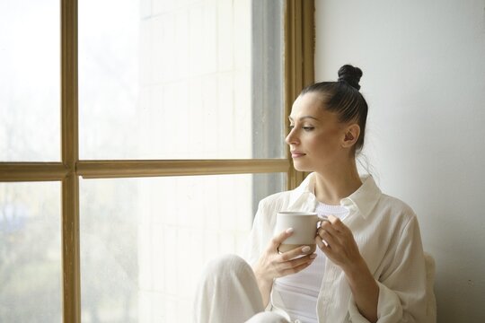 Calm and peaceful woman in white shirt sitting on the windowsill with a cup of tea or coffee in her hands