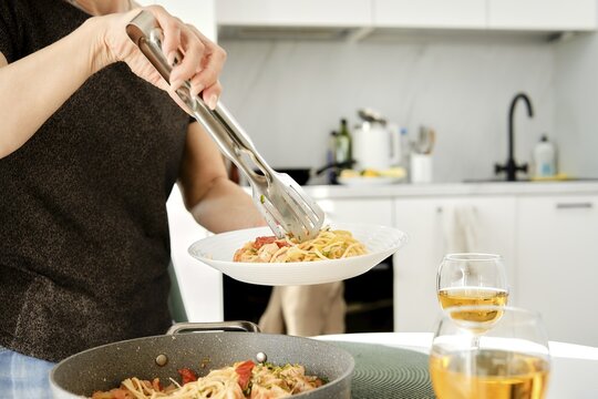 A person uses tongs to serve pasta onto a white plate from a pan in a contemporary kitchen. Two glasses of drink sit on the counter, adding to the inviting atmosphere of the home