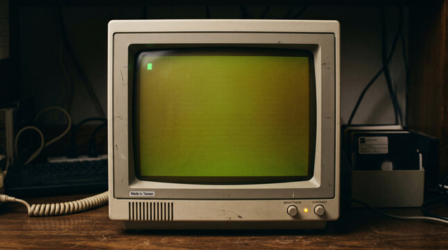 A vintage beige computer monitor with a glowing green screen and manual brightness and contrast knobs sits prominently on a wooden table in soft lighting.