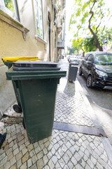 Naklejka premium Green and yellow trash bins positioned on a cobblestone sidewalk beside a parked black car and a building with windows in a sunny urban environment