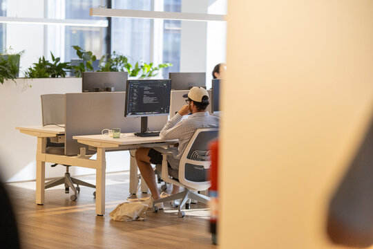Individual seated at a modern office desk working on a computer with programming code displayed on the monitor, plants and colleagues visible in the background