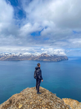 Lonely man standing on mountain cliff overlooking ocean and snowy peaks, Unalaska, Alaska. Hiker admiring sea and snow-covered mountains in USA. 