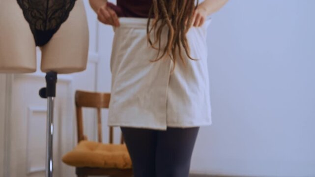 A dreadlocked seamstress admires her handmade skirt in a full-length studio mirror, smoothing the fabric over her hips. She lifts the hem slightly and checks the evenness of the stitches