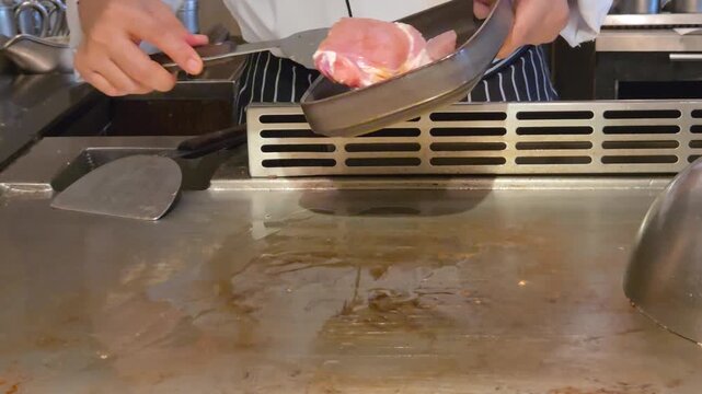 Eye Level Close Up View of Professional Teppanyaki Chef Using a Spatula to Place a Raw Kurobuta (Kagoshima Berkshire) Pork Meat Steak on a Hot Flat Griddle