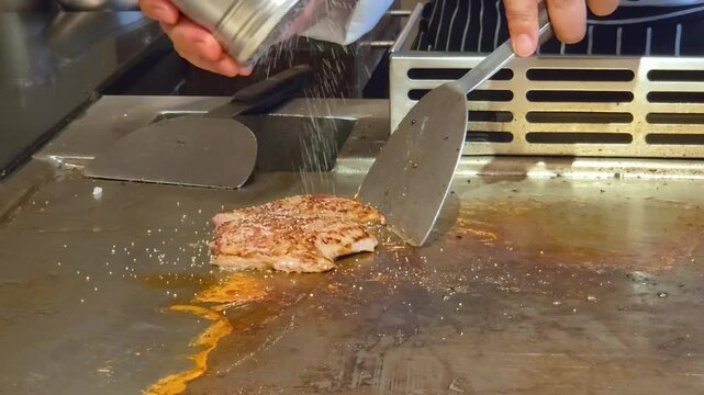 Eye Level Close Up View of Professional Teppanyaki Chef Seasoning Meat with a Stainless Steel Shaker and Slicing a Cooked Steak with a Knife on a Hot Flat Griddle