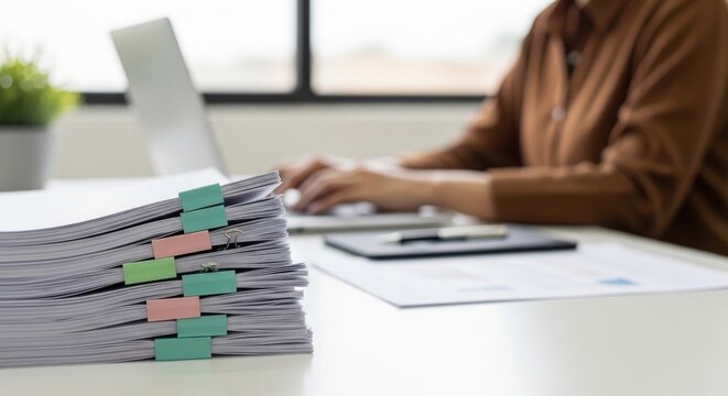 Stack of documents in binders on desk with person working on laptop in blurred background at bright office for administrative work and organization concept