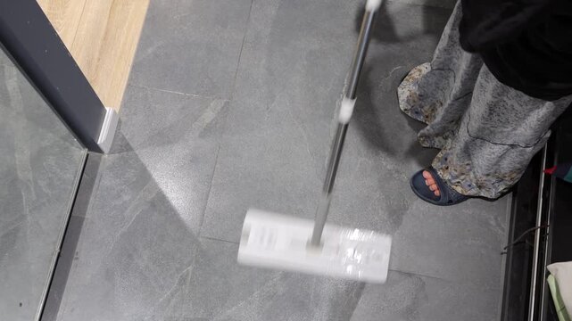 A teenager guides a flat mop over gray tile near a wooden floor threshold during entryway cleaning.