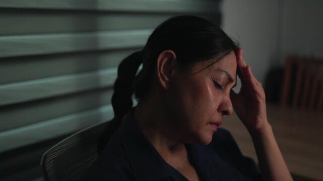 Overwhelmed by Pressure: A woman in professional attire clutches her head, eyes closed in a moment of deep stress and contemplation. The dimly lit room with blinds suggests a private.