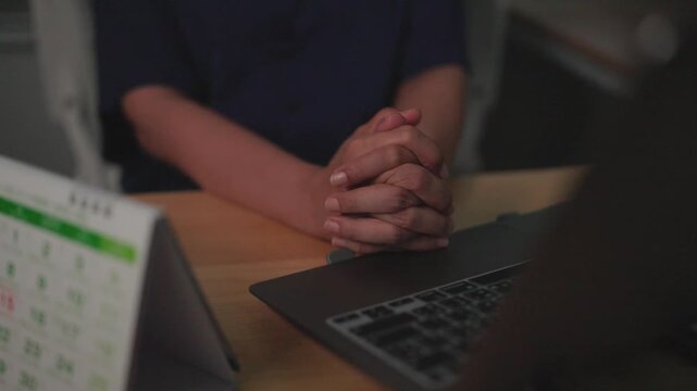 Anxious Waiting: Clasped hands rest on a desk beside a laptop and a desk calendar, embodying a moment of anticipation and perhaps unease, possibly during a meeting or waiting for important news.