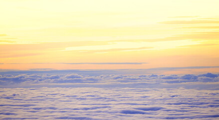 Fototapeta premium This aerial landscape presents a thick blanket of clouds spanning the horizon under a soft sunset sky featuring warm yellow and orange hues that transition to gentle purple tones above the clouds