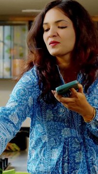 Young beautiful Indian Asian woman cooking food in a pan in the kitchen while speaking on a speakerphone, multitasking and managing kitchen tasks efficiently