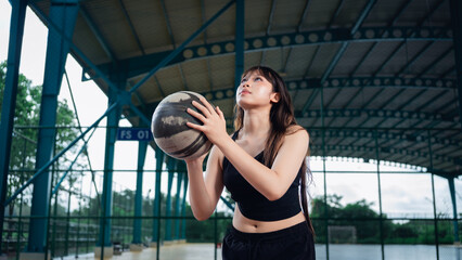 Naklejka premium Young woman in athletic wear holding basketball on outdoor sports court during golden hour, preparing for exercise or game, with a covered roof structure and greenery in background