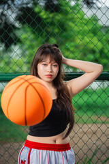 Naklejka premium Young woman with long hair and casual attire posing on a basketball court near a chain-link fence with a orange basketball in front of her and lush green trees background
