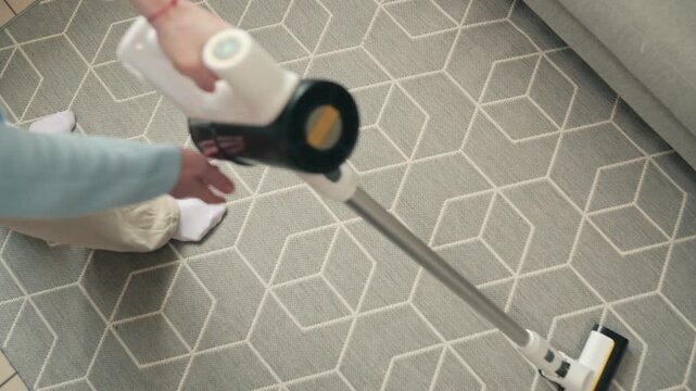 Young woman using cordless vacuum cleaner to clean carpet in living room during regular household cleaning routine at home
