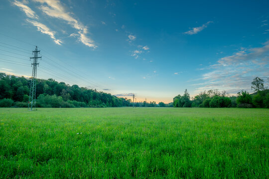 Expansive wide open green field in the tranquil countryside featuring distant power transmission towers and a thick forest edge under a magnificent blue sky with gentle sunset colors.