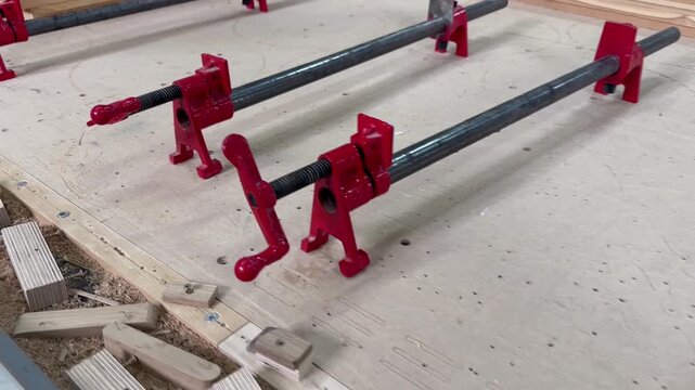 Woodworker's tools rest on rough sawdust floor. Red clamps hold wood in place during assembly. Blue pencil lies nearby, used for marking measurements
