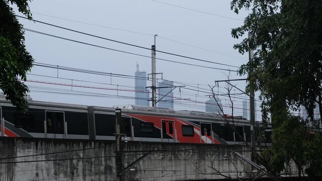 Modern commuter train traveling on elevated railway bridge with overhead wires, concrete barrier, and urban transportation infrastructure.