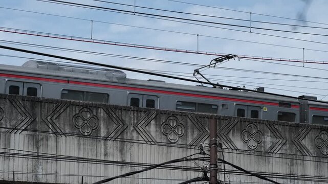 Modern commuter train traveling on elevated railway bridge with overhead wires, concrete barrier, and urban transportation infrastructure.