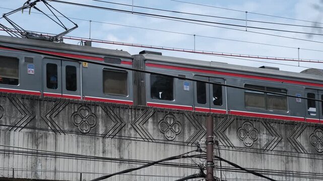 Modern commuter train traveling on elevated railway bridge with overhead wires, concrete barrier, and urban transportation infrastructure.