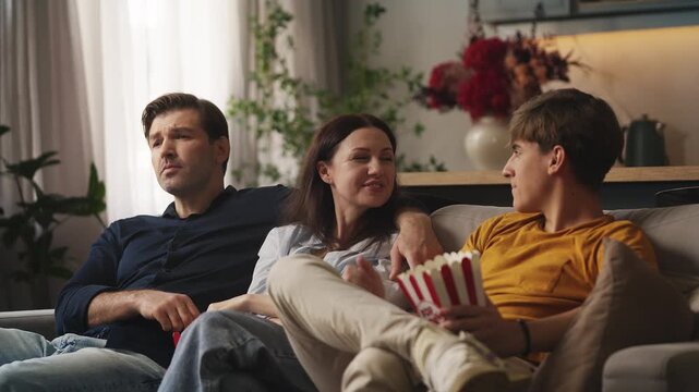 Cheerful adolescent boy and his parents watching a movie and eating popcorn