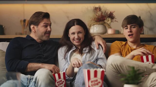 Happy parents and their teen son eating popcorn while watching movie at home