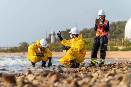 Hazmat workers in yellow suits test ocean water samples. An ecologist records data on a digital tablet. Petrochemical storage tanks stand behind the scene