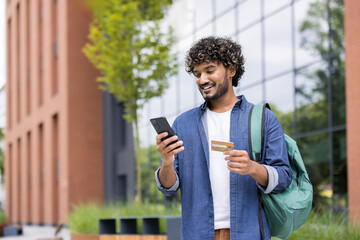Obraz na płótnie Canvas Young indian man with curly hair and backpack smiling, making an online purchase using his smartphone and a credit card while standing on a modern urban campus