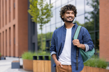 Obraz premium Smiling young indian student with curly hair confidently walking on a modern university campus, carrying a backpack and looking away, representing higher education, success, and a bright future