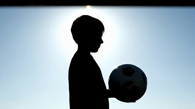 Silhouette of a boy holding a soccer ball standing against a bright sun and blue sky.