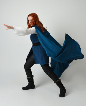 full length portrait redhead female model wearing medieval fantasy warrior historical costume with blue hood cloak. walking away with magical spell arm gesture, silhouette figure on studio background.