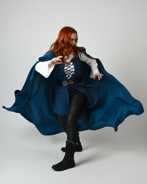 full length portrait redhead female model wearing medieval fantasy warrior historical costume with blue hood cloak. walking away with magical spell arm gesture, silhouette figure on studio background.