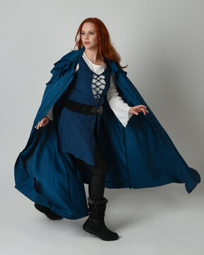 full length portrait redhead female model wearing medieval fantasy warrior historical costume with blue hood cloak. walking away with magical spell arm gesture, silhouette figure on studio background.