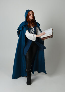 full length portrait of female model wearing medieval fantasy warrior historical costume with blue cloak. silhouette figure,  holding magical spell book on studio background.
