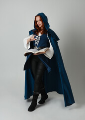 Naklejka premium full length portrait of female model wearing medieval fantasy warrior historical costume with blue cloak. silhouette figure, holding magical spell book on studio background. 