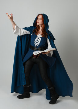 full length portrait of female model wearing medieval fantasy warrior historical costume with blue cloak. silhouette figure,  holding magical spell book on studio background.

