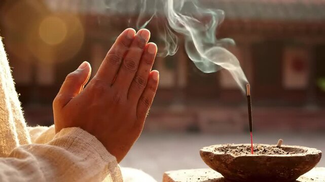 Hands in prayer pose next to burning incense stick, smoke rising, spiritual meditation, tranquil atmosphere, soft sunlight