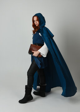 full length portrait of female model wearing medieval fantasy warrior historical costume with blue cloak. silhouette figure,  holding magical spell book on studio background.
