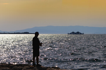 Fototapeta premium Silhouette of a fisherman with a fishing rod standing on the rocky shore during sunset, with a passenger ferry boat and mountains on the shimmering sea in the background.