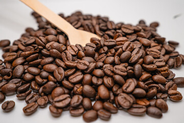 Obraz premium Roasted coffee beans piled on a white surface with a wooden spoon, representing fresh coffee ingredients and cafe culture