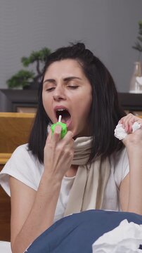 Upset ill young brunette lying in bedroom bed and coughing and using throat sprey while is treated at home during flu sickness