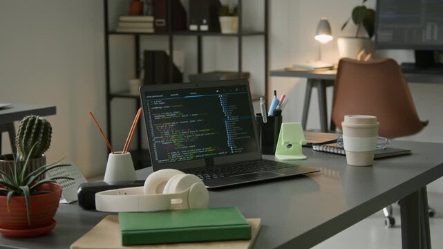 No people medium shot of software developer desk in office, with laptop computer displaying code lines on black screen, headphones, planner, coffee cup, pot plants, aroma sticks, before working day