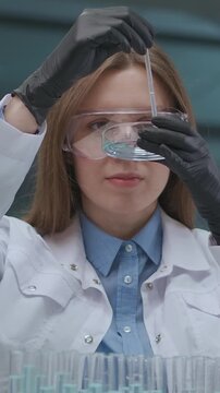 Focused female scientist wearing safety goggles and lab coat conducting chemical experiments with pipette and petri dish in a laboratory setting surrounded by multiple test tubes