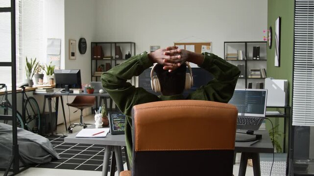 Medium rear shot of unrecognizable black male developer reclining in chair and stretching, after running vibe coding agent on computer, controlling execution of prompt for web design project
