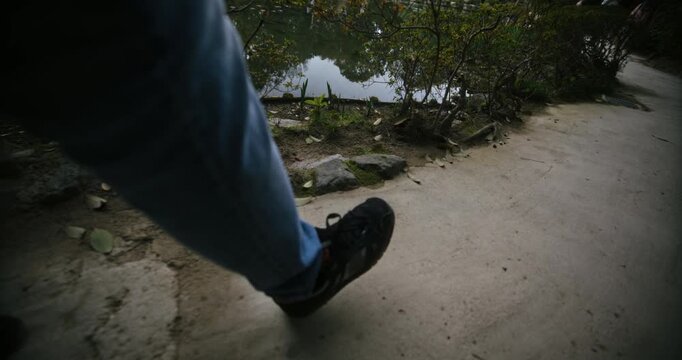 Low shot on person's legs as they walk on concrete near grass and pond
