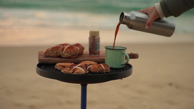 Coastal Morning Brew: A rustic outdoor scene on a sandy beach features freshly baked bread rolls and a warm drink being poured from a thermos into a mug.