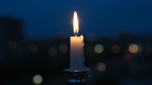 A single candle flame in the dark against the bokeh of city lights. Wax has dripped down the side