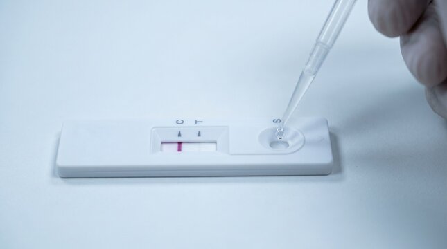 A hand drops liquid onto a rapid diagnostic test kit on a white surface with a red indicator line visible.