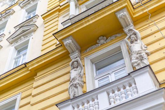 Ornate historical building facade with yellow stucco, white balcony balustrade, and classical caryatid statue sculptures in Prague, Czech Republic.